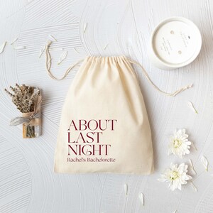 May include: A beige cotton drawstring pouch featuring the dark red text "ABOUT LAST NIGHT" and "Rachel's Bachelorette" printed on the front. The bag is accompanied by a small bundle of dried lavender, a white candle, and white flower petals.