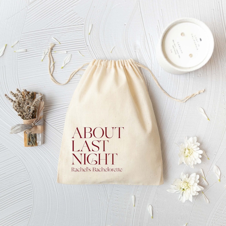 May include: A beige cotton drawstring pouch featuring the dark red text "ABOUT LAST NIGHT" and "Rachel's Bachelorette" printed on the front. The bag is accompanied by a small bundle of dried lavender, a white candle, and white flower petals.