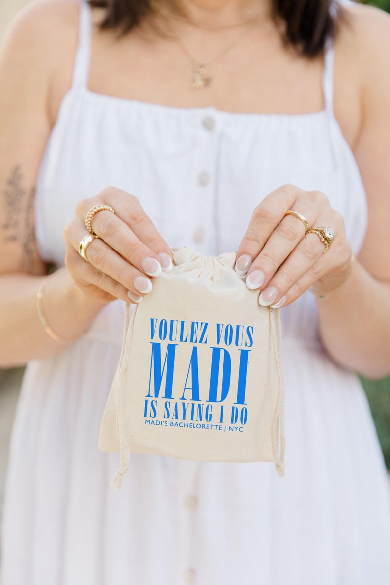 May include: A white drawstring bag with the text "Voulez Vous Madi Is Saying I Do" in blue lettering. The bag is decorated with the text "Madi's Bachelorette | NYC" in smaller blue lettering.