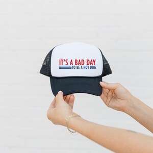 May include: A black and white trucker hat with the text "IT'S A BAD DAY TO BE A HOT DOG" in red and blue. The hat has a white front panel and a black brim and mesh back.