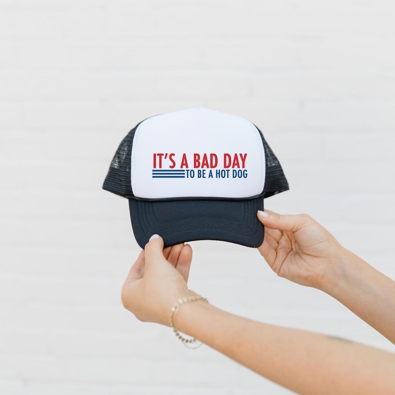 May include: A black and white trucker hat with the text "IT'S A BAD DAY TO BE A HOT DOG" in red and blue. The hat has a white front panel and a black brim and mesh back.