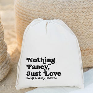 May include: A white drawstring bag with the text "Nothing Fancy, Just Love" in a bold, retro font. Below the text is "Bolaji & Molly | 10.12.24". The bag is on a sandy surface with a woven basket in the background.