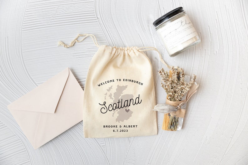Pode incluir: Uma bolsa de algod&atilde;o branco com cord&atilde;o de ajuste com um mapa da Esc&oacute;cia e o texto "Bem-vindos a Edimburgo, Esc&oacute;cia, Brooke & Albert 6.7.2023". A bolsa est&aacute; ao lado de um pequeno buqu&ecirc; de lavanda seca e um pote de vela de vidro transparente com tampa preta.