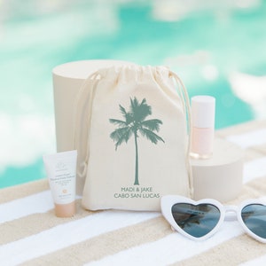 May include: A white canvas bag with a green palm tree design and the text "Madi & Jake Cabo San Lucas". The bag is sitting on a white and brown striped towel with a white heart-shaped sunglasses, a small bottle of pink nail polish, and a small tube of hand cream.