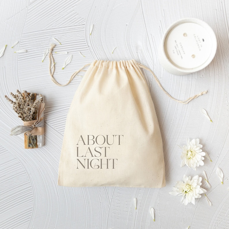 May include: A beige cotton drawstring pouch with the text ABOUT LAST NIGHT printed in dark gray. Beside the pouch is a bundle of dried lavender and sage tied with a ribbon, a white candle in a round container, and two white flowers.