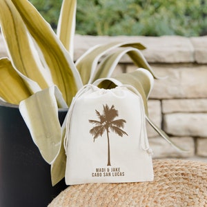 May include: A white cotton drawstring bag with a brown palm tree design and the text "Madi & Jake Cabo San Lucas" printed on it.