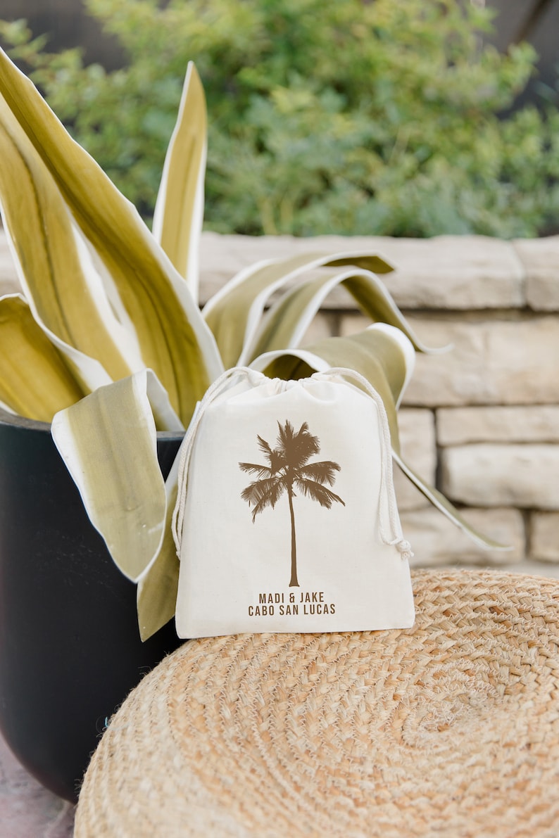 May include: A white cotton drawstring bag with a brown palm tree design and the text "Madi & Jake Cabo San Lucas" printed on it.