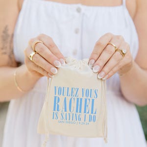 May include: A white drawstring bag with the text "Voulez vous Rachel is saying I do San Diego | 9.29.24" printed in blue.