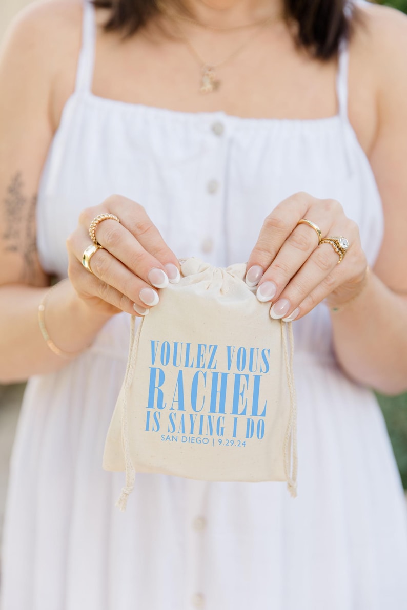 May include: A white drawstring bag with the text "Voulez vous Rachel is saying I do San Diego | 9.29.24" printed in blue.