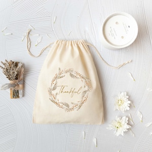 May include: A white cotton drawstring bag with a gold floral wreath and the word "Thankful" printed on it. The bag is surrounded by white flowers and a white candle.