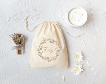 May include: A white cotton drawstring bag with a gold floral wreath and the word "Thankful" printed on it. The bag is surrounded by white flowers and a white candle.