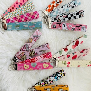 Könnte beinhalten: Eine Sammlung von Schlüsselanhängern mit verschiedenen Mustern, darunter rosa Makronen, Fische und Halloween-Designs. Jeder Schlüsselanhänger hat einen silberfarbenen Verschluss und ein gemustertes Stoffband.