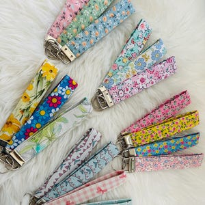 Op de afbeelding: Een verzameling stoffen pols keychains in diverse bloemen- en patroonontwerpen. Keychains hebben een metalen sluiting en ring. Kleuren zijn roze, geel, blauw, groen en wit. De keychains liggen op een witte, zachte ondergrond.