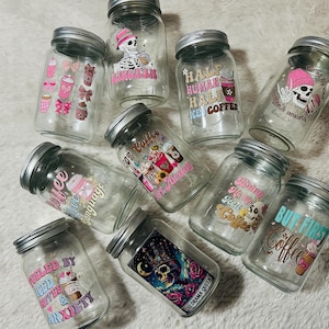 Peut inclure: Plusieurs bocaux en verre transparent avec des couvercles argentés, ornés d'autocollants colorés. Les autocollants présentent des graphiques sur le thème du café, des crânes et des phrases comme "But First Coffee" et "Fueled by Iced Coffee & Anxiety."