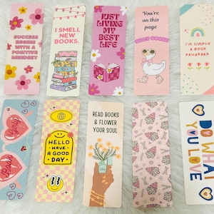 May include: A selection of vibrant bookmarks with diverse designs and text. The bookmarks showcase floral patterns, motivational quotes, and charming illustrations. Some include phrases such as "I Smell New Books" and "Do What You Love."