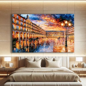 May include: A large rectangular canvas wall art piece depicting a vibrant city square at sunset. The painting features warm orange and yellow sky tones, illuminated building facades with arched walkways, and a tall street lamp reflecting on wet ground.