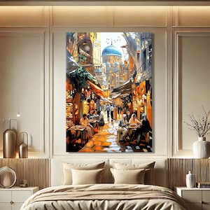 May include: A large framed canvas print showing a bustling street scene with a blue-domed building. The artwork features people dining at tables, with warm orange and yellow tones. The print is displayed above a bed in a bedroom setting.