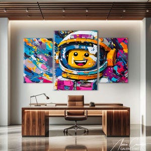 May include: A three-panel artwork featuring a Lego astronaut with a smiling face, set against a vibrant, abstract background of colorful paint splatters. The astronaut's helmet is blue, yellow, and white. The artwork is displayed above a wooden desk.