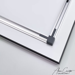 May include: A close-up of a white framed art piece with a silver metal frame. The corners of the frame are connected with black plastic corner pieces. The bottom right corner of the art piece has a signature and the words "GALERIE D'ART".