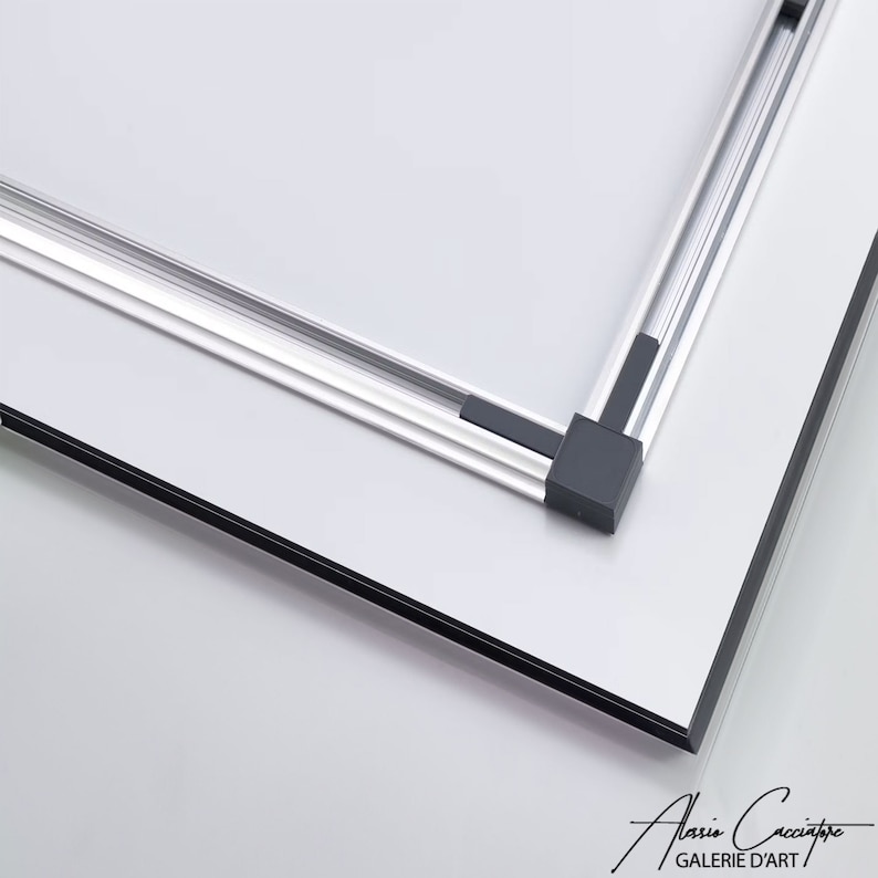 May include: A close-up of a white framed art piece with a silver metal frame. The corners of the frame are connected with black plastic corner pieces. The bottom right corner of the art piece has a signature and the words "GALERIE D'ART".
