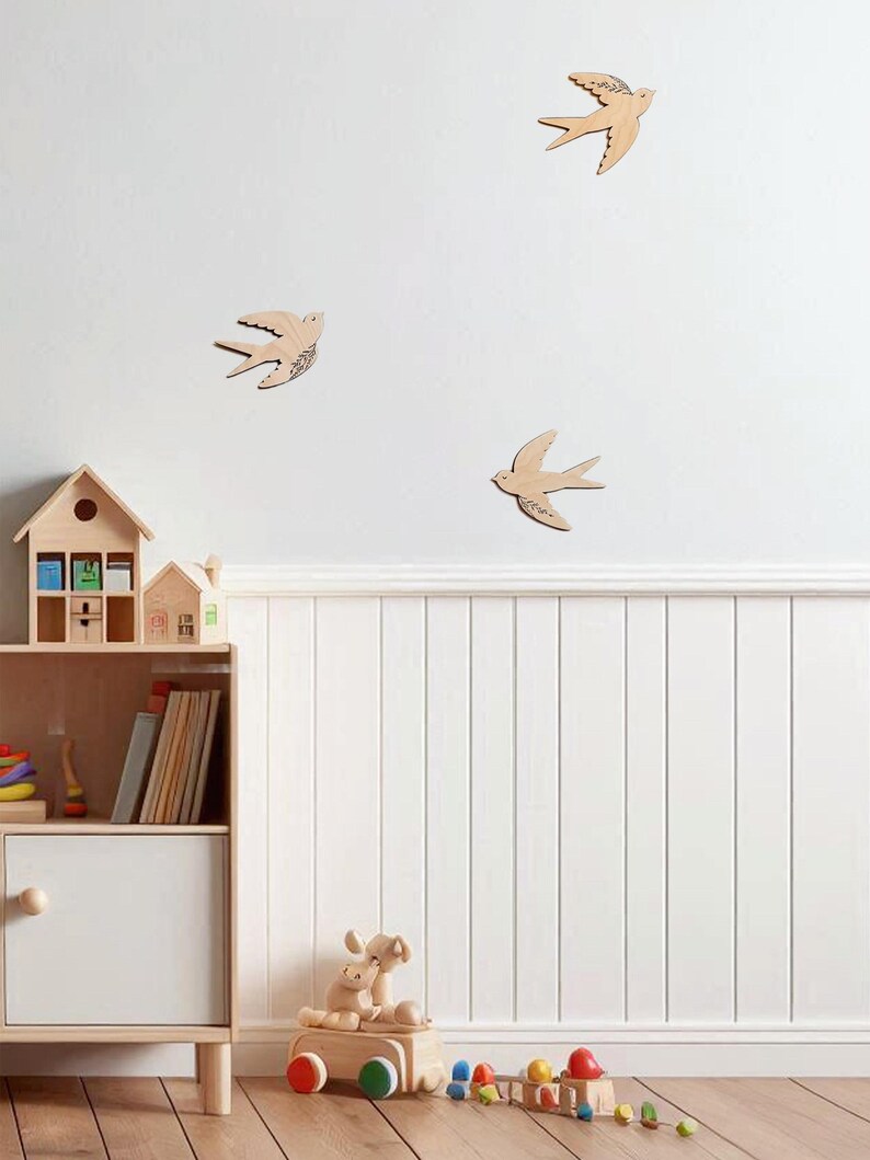 Puede incluir: Decoraci&oacute;n de pared de madera con forma de golondrinas de varios tama&ntilde;os, montada en una pared blanca. La habitaci&oacute;n tambi&eacute;n cuenta con una estanter&iacute;a de madera con juguetes y libros, y un tren de juguete de madera. El tema general es una habitaci&oacute;n infantil.
