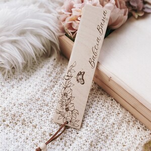Op de afbeelding: Een houten bladwijzer met de tekst "Babcia Basia" gegraveerd in een cursief lettertype. De bladwijzer heeft een bloemen- en vlinderontwerp, met een bruin leren koord en een houten kraal. De achtergrond omvat bloemen en een wit gebreid oppervlak.