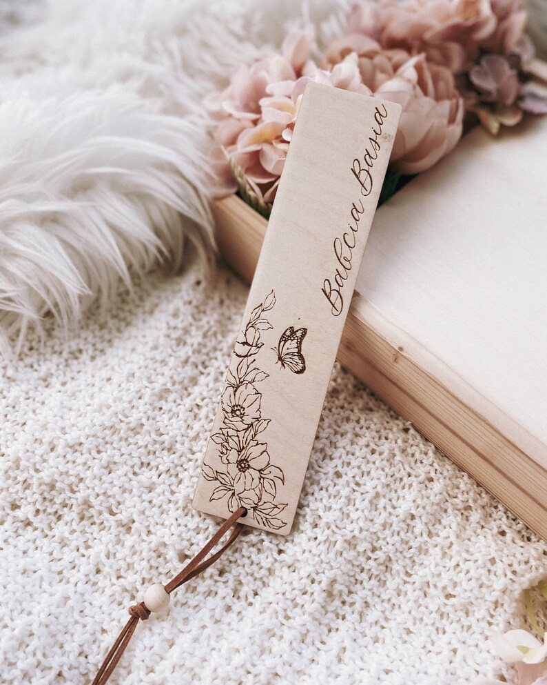 Op de afbeelding: Een houten bladwijzer met de tekst "Babcia Basia" gegraveerd in een cursief lettertype. De bladwijzer heeft een bloemen- en vlinderontwerp, met een bruin leren koord en een houten kraal. De achtergrond omvat bloemen en een wit gebreid oppervlak.