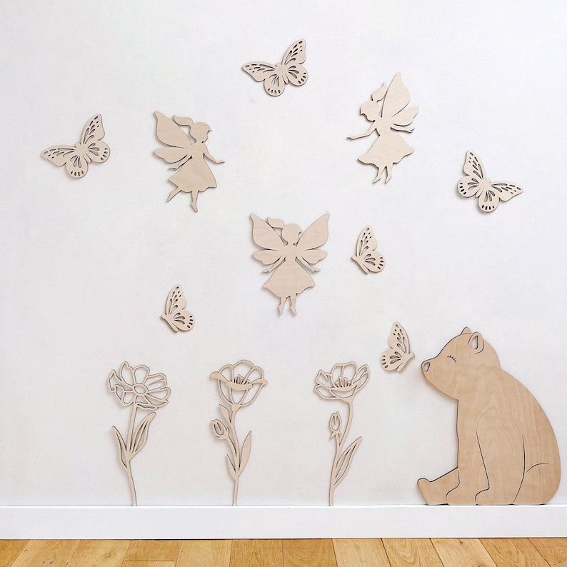 May include: Wooden wall decor featuring a bear, flowers, butterflies, and fairies. The bear is sitting and looking up at a butterfly. The flowers are simple and have a single bloom. The butterflies are in various positions and sizes. The fairies are in a flying pose with wings outstretched.