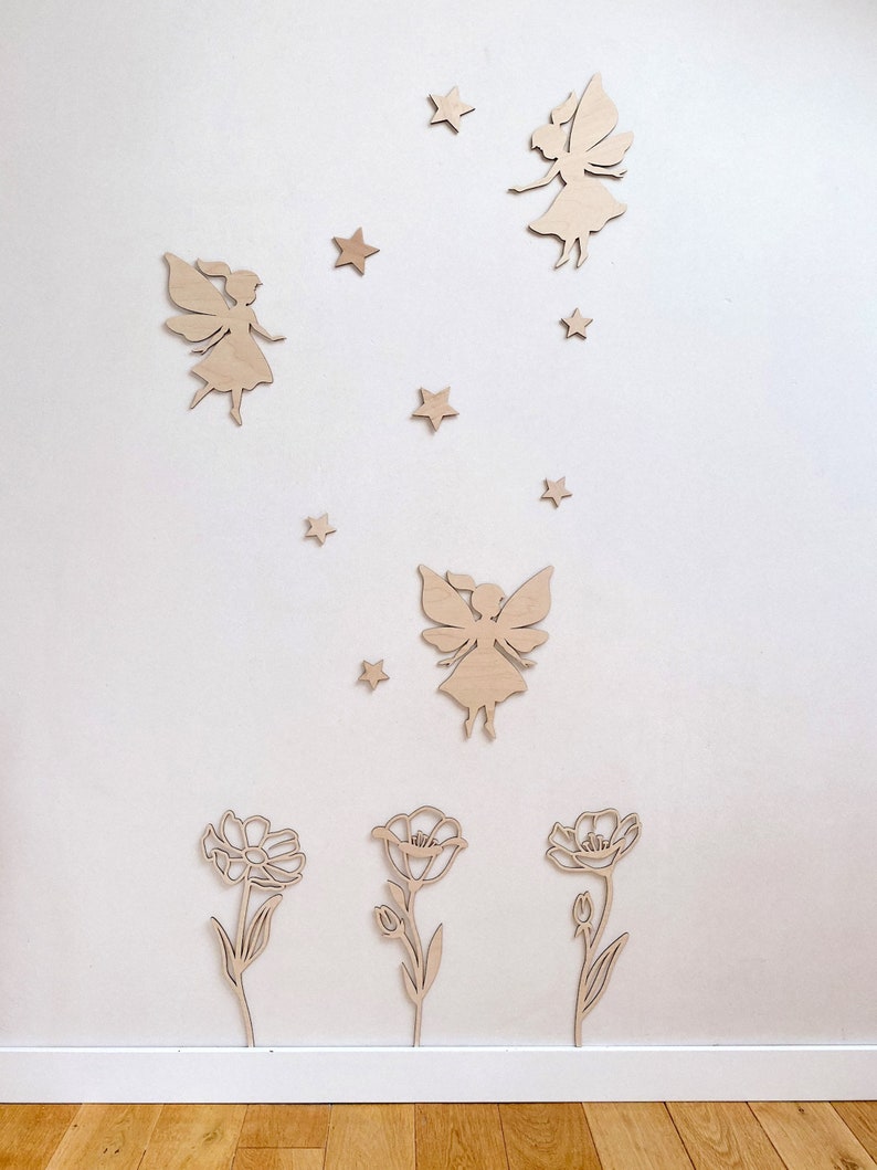 May include: Wooden wall decor featuring three fairies and three flowers. The fairies are flying with their wings spread out. The flowers are simple and have a stem and leaves. The decor is painted in a light wood color.