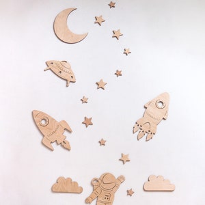 May include: Wooden wall decor featuring a crescent moon, stars, a rocket, a spaceship, a cloud, and an astronaut. The decor is painted in shades of brown.