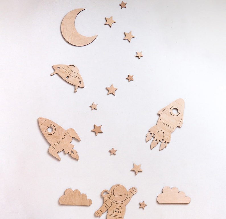 May include: Wooden wall decor featuring a crescent moon, stars, a rocket, a spaceship, a cloud, and an astronaut. The decor is painted in shades of brown.