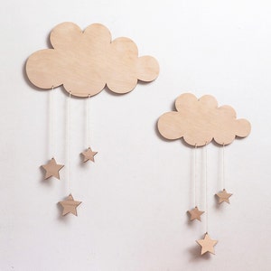 Puede incluir: Dos formas de nube de madera con estrellas colgando de ellas. Las nubes son de color marrón claro y las estrellas son de un marrón ligeramente más oscuro. Las nubes están colgadas en una pared blanca.