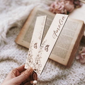 Op de afbeelding: Twee houten bladwijzers met bloemen- en vlindermotieven. E&eacute;n bladwijzer heeft de naam "Babcia Basia" en de andere "Amelia" gegraveerd in een sierlijk lettertype. Elke bladwijzer heeft een leren kwast met houten kralen.