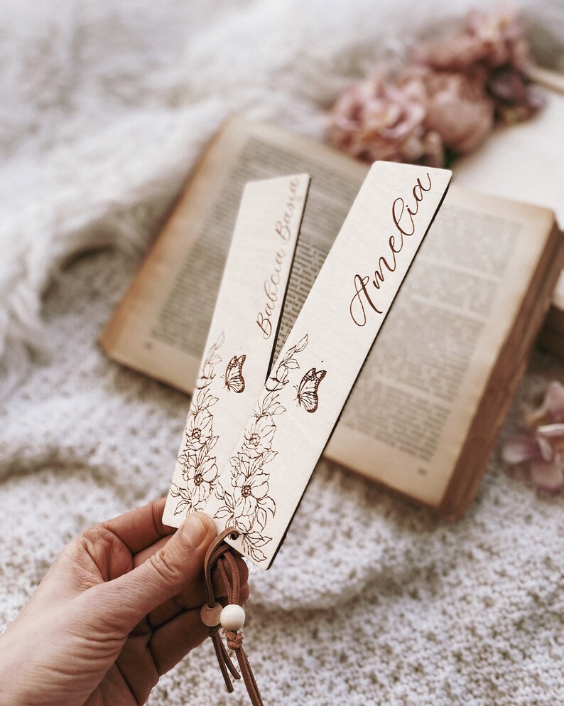 Op de afbeelding: Twee houten bladwijzers met bloemen- en vlindermotieven. E&eacute;n bladwijzer heeft de naam "Babcia Basia" en de andere "Amelia" gegraveerd in een sierlijk lettertype. Elke bladwijzer heeft een leren kwast met houten kralen.