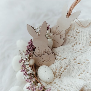 May include: A white wicker basket filled with white speckled eggs and pink flowers. Two wooden bunny-shaped Easter tags are attached to the basket, one with the name "Emil" engraved on it.