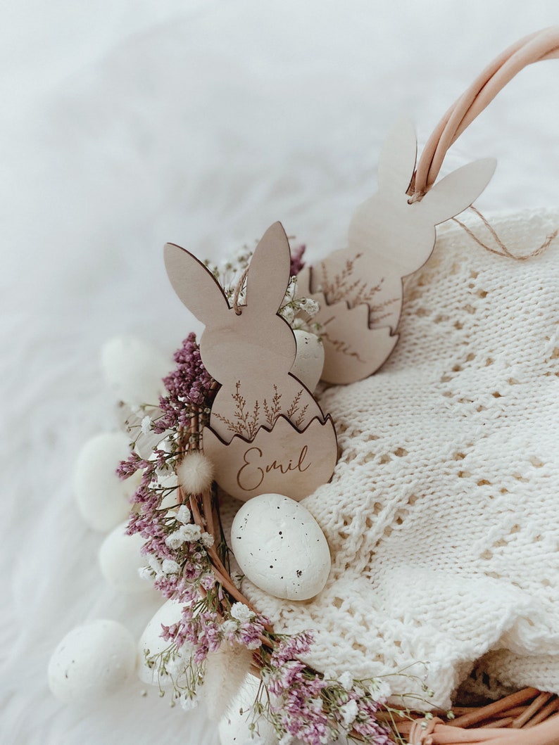 May include: A white wicker basket filled with white speckled eggs and pink flowers. Two wooden bunny-shaped Easter tags are attached to the basket, one with the name "Emil" engraved on it.