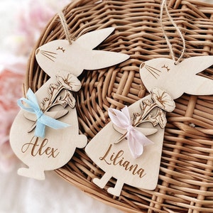 May include: Two wooden bunny ornaments with floral designs and personalised names, Alex and Liliana. The bunnies are holding blue and pink ribbons.