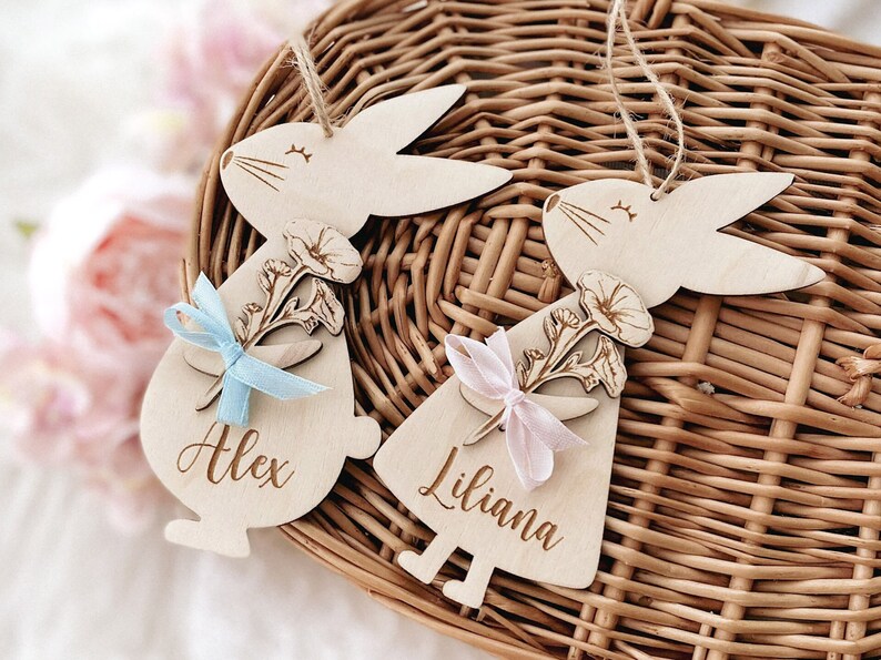 May include: Two wooden bunny ornaments with floral designs and personalised names, Alex and Liliana. The bunnies are holding blue and pink ribbons.