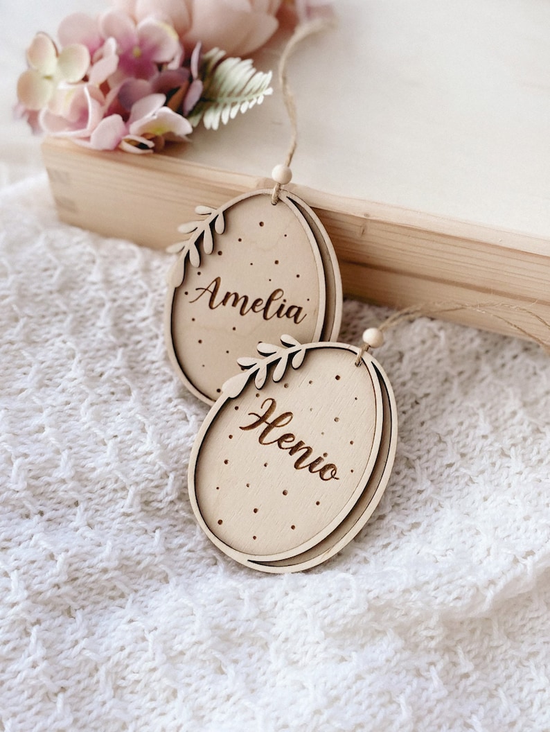 Peut inclure: Deux d&eacute;corations d'&oelig;ufs de P&acirc;ques en bois avec des cintres en ficelle. Les &oelig;ufs sont d&eacute;cor&eacute;s de points blancs et de brindilles feuillues. L'&oelig;uf du haut est grav&eacute; du nom "Amelia" et l'&oelig;uf du bas est grav&eacute; du nom "Henio".