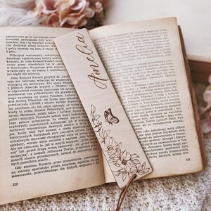 Op de afbeelding: Houten bladwijzer met de naam "Amelia" in sierlijke letters. Het heeft een bloemen- en vlinderontwerp, een leren koord en een houten kraal. De bladwijzer ligt in een open boek.