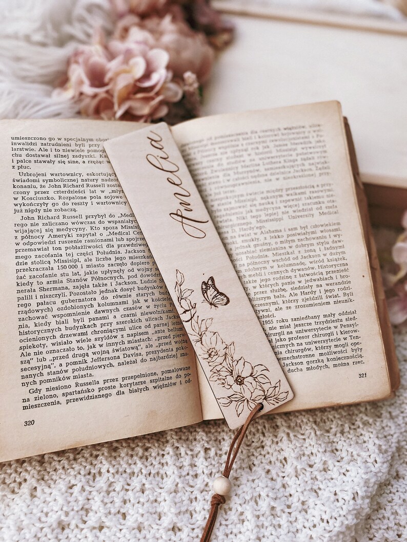 Op de afbeelding: Houten bladwijzer met de naam "Amelia" in sierlijke letters. Het heeft een bloemen- en vlinderontwerp, een leren koord en een houten kraal. De bladwijzer ligt in een open boek.