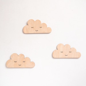 May include: Three wooden clouds with painted eyes and smiles. The clouds are light brown and have a simple, whimsical design.