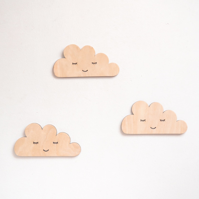 May include: Three wooden clouds with painted eyes and smiles. The clouds are light brown and have a simple, whimsical design.