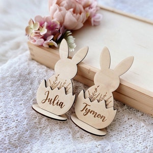 May include: Two wooden bunny place card holders with the names Julia and Tymon engraved on them. The bunnies are sitting in egg-shaped nests with a floral design.