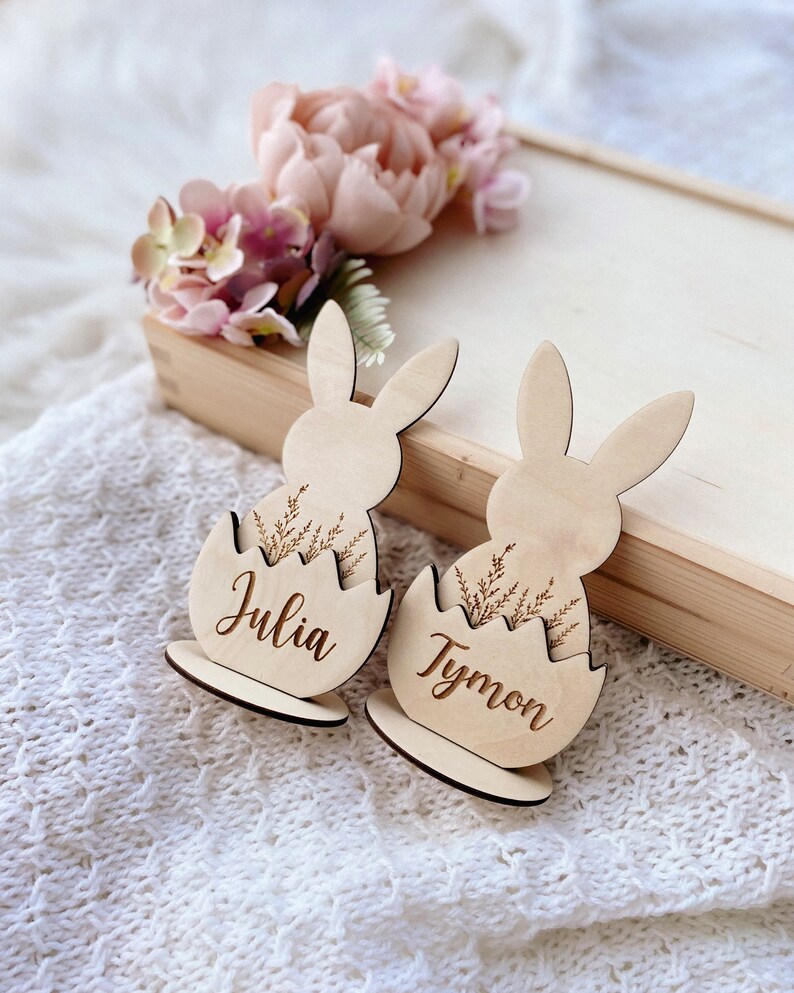 May include: Two wooden bunny place card holders with the names Julia and Tymon engraved on them. The bunnies are sitting in egg-shaped nests with a floral design.