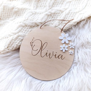 Peut inclure: Une pancarte ronde en bois avec le nom "Olivia" grav&eacute; en &eacute;criture cursive. La pancarte est d&eacute;cor&eacute;e de fleurs en feutre blanc.