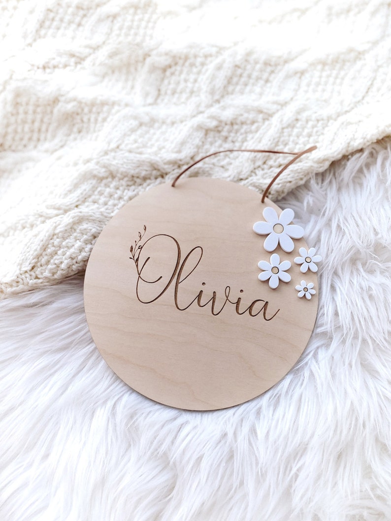 Peut inclure: Une pancarte ronde en bois avec le nom "Olivia" grav&eacute; en &eacute;criture cursive. La pancarte est d&eacute;cor&eacute;e de fleurs en feutre blanc.