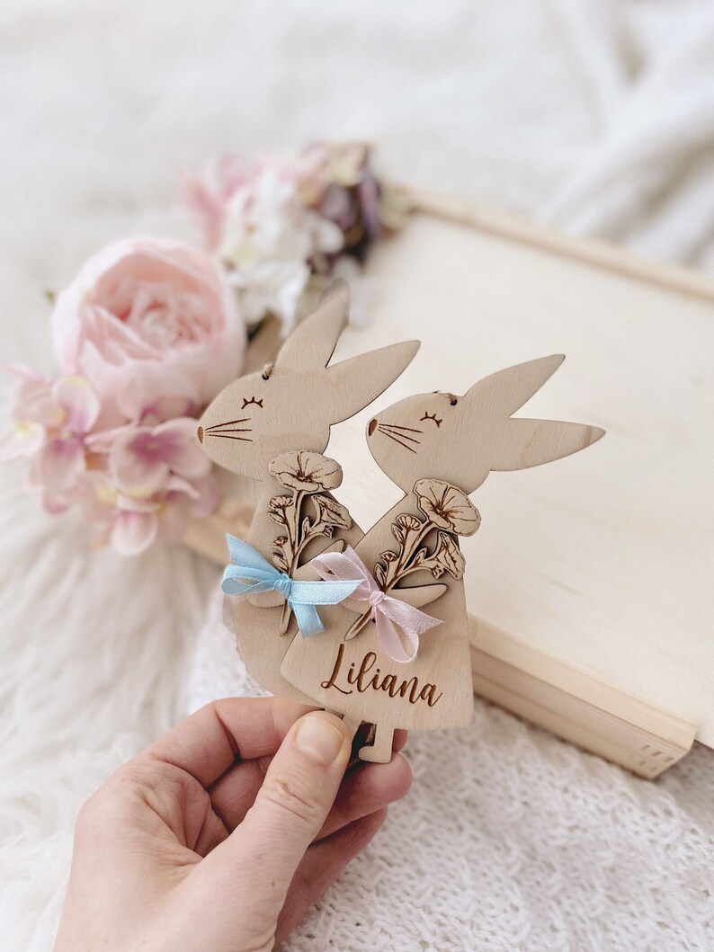 May include: Two wooden bunny ornaments with floral details and blue and pink ribbons. The bunny on the right has the name "Liliana" engraved on it.
