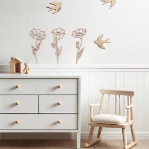 Puede incluir: Escena de guarder&iacute;a con decoraci&oacute;n de pared de madera de p&aacute;jaros y flores. Una c&oacute;moda blanca con pomos de madera y una peque&ntilde;a mecedora de madera en primer plano. Una peque&ntilde;a casa de madera y un oso de peluche est&aacute;n en la c&oacute;moda.