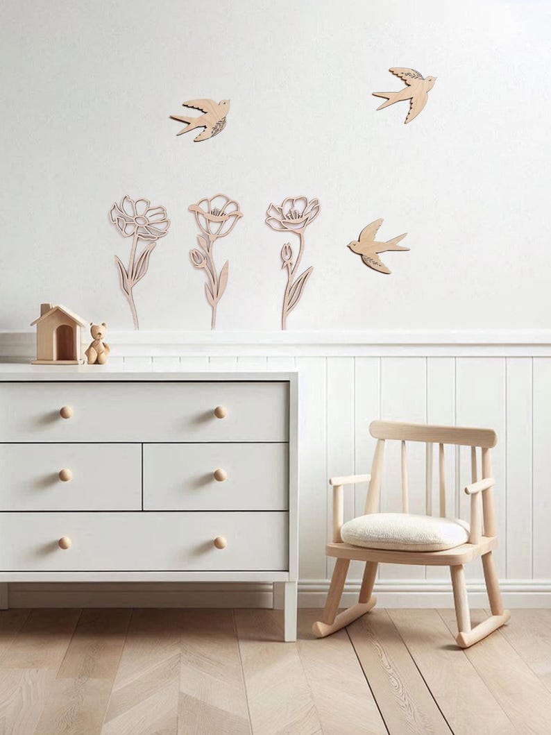 Puede incluir: Escena de guarder&iacute;a con decoraci&oacute;n de pared de madera de p&aacute;jaros y flores. Una c&oacute;moda blanca con pomos de madera y una peque&ntilde;a mecedora de madera en primer plano. Una peque&ntilde;a casa de madera y un oso de peluche est&aacute;n en la c&oacute;moda.